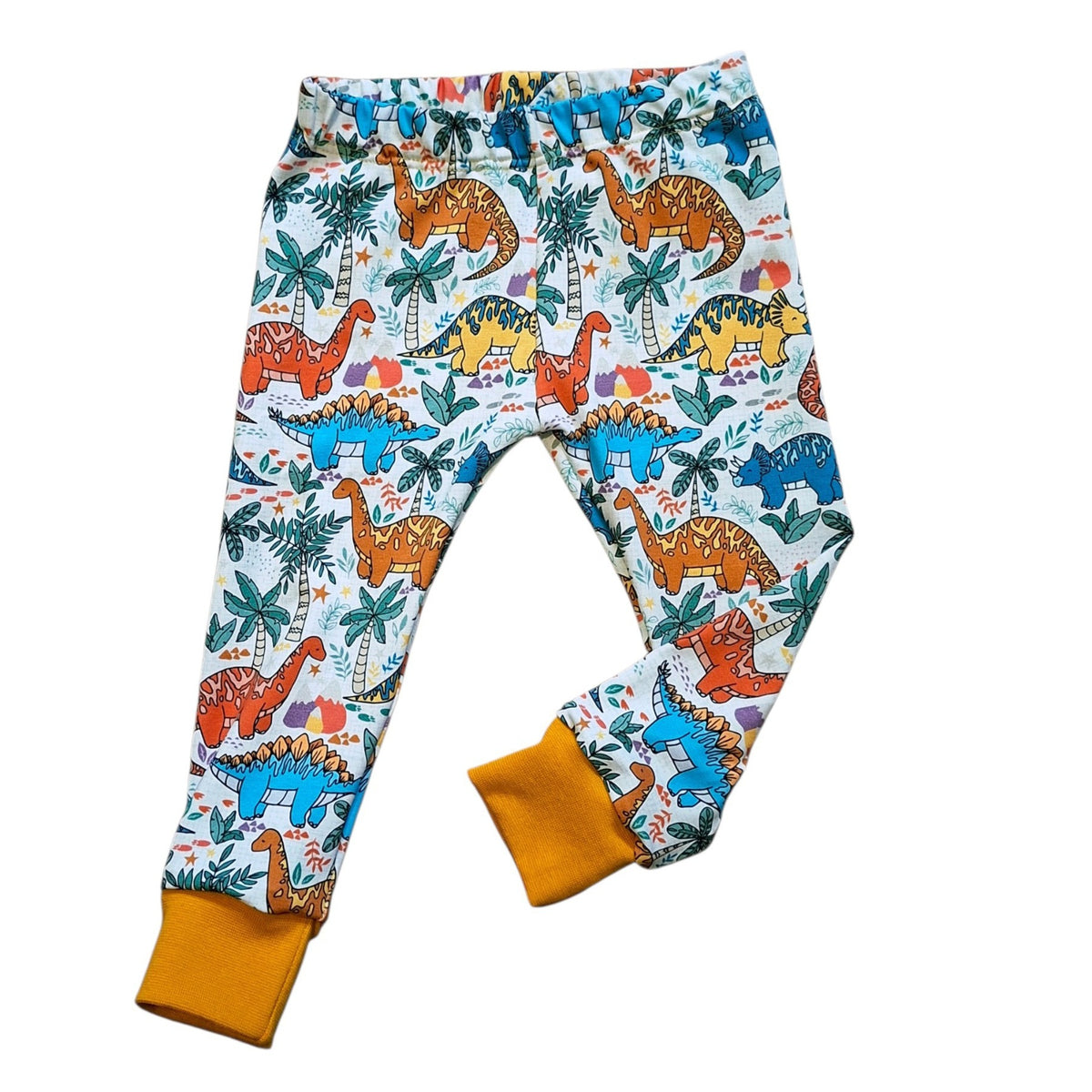 A pair of children's leggings featuring an all-over colorful dinosaur and palm tree print on a white background, with solid mustard yellow cuffs.