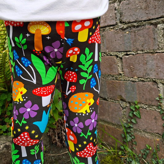 Colorful patterned leggings with mushrooms, flowers, and hedgehogs worn by a person against a brick wall.