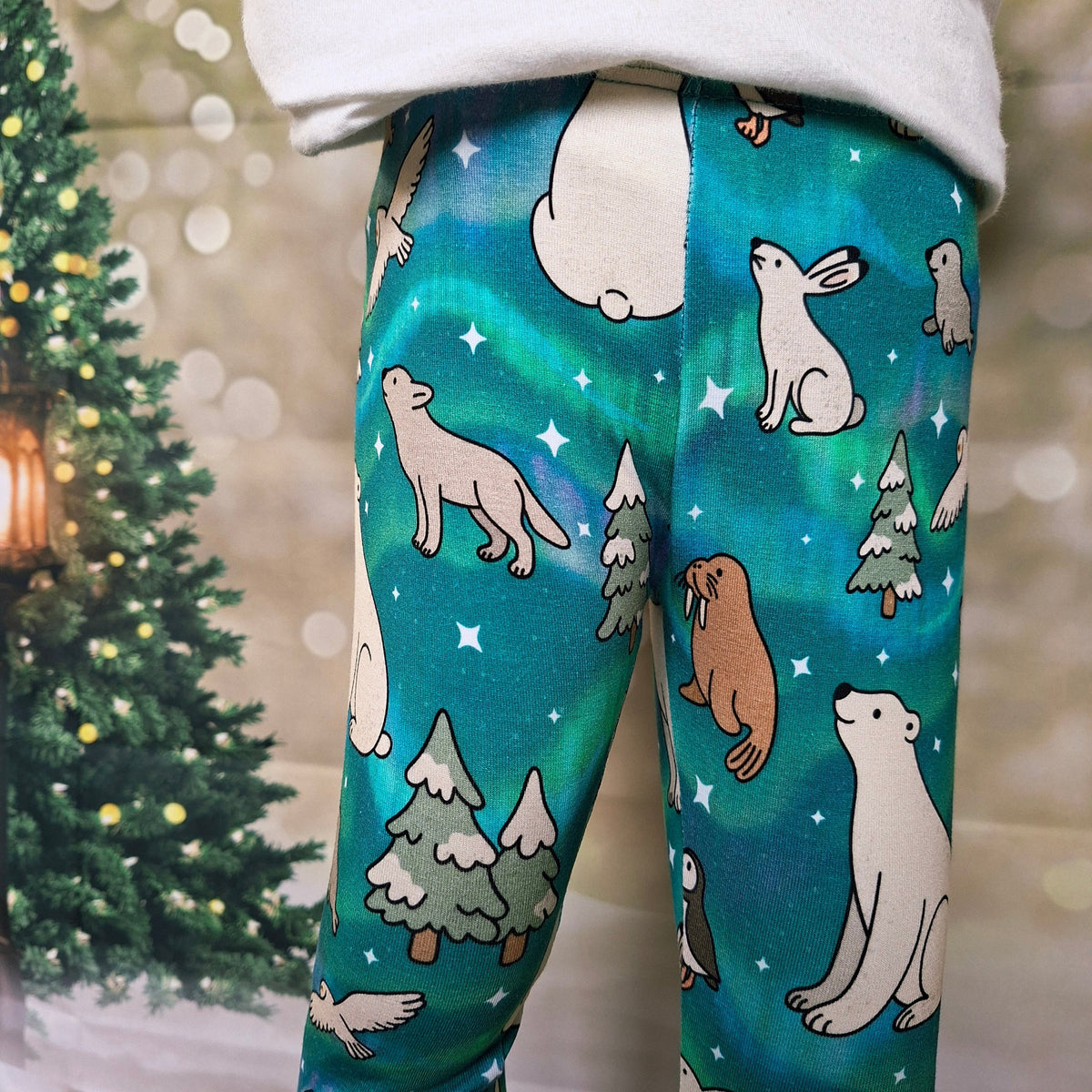 A child wearing teal and green patterned leggings with a winter animal print, featuring polar bears, wolves, rabbits, and snowy trees. The background shows a blurred Christmas tree and warm bokeh lights.