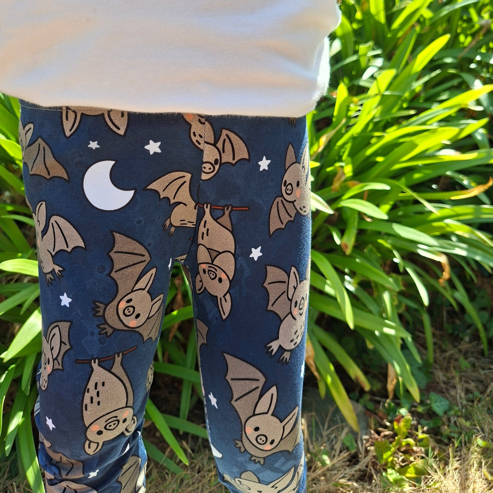 A child outdoors wearing dark navy leggings with a cartoon bat print, a white top, standing in front of green plants.