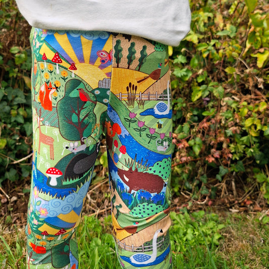 A child wearing colorful, whimsical landscape-print leggings and a white sweatshirt, standing outdoors in front of green foliage.