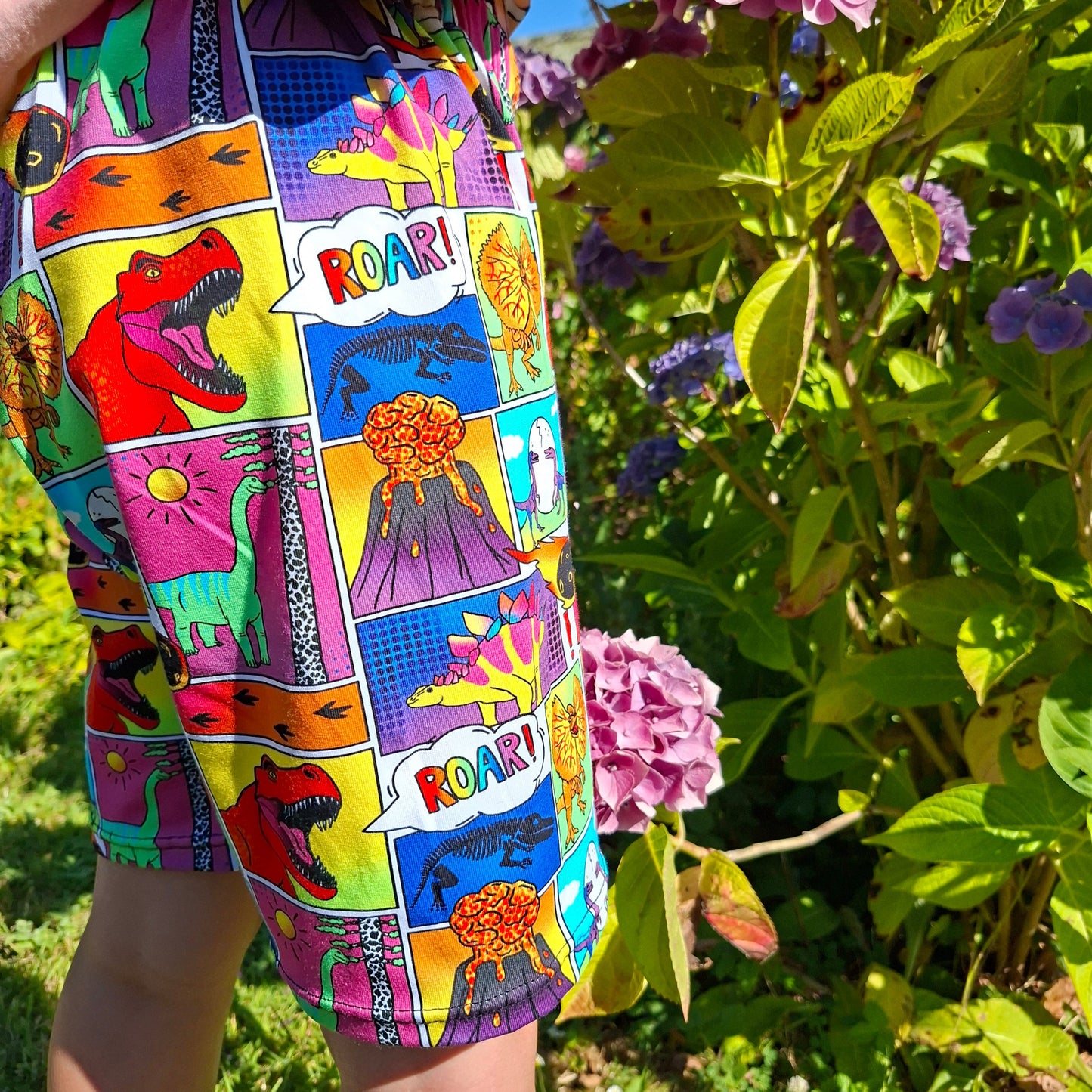 A close-up of a colourful children's shorts or skirt featuring a comic-book style dinosaur print, worn by a child outdoors in front of green hydrangea bushes.