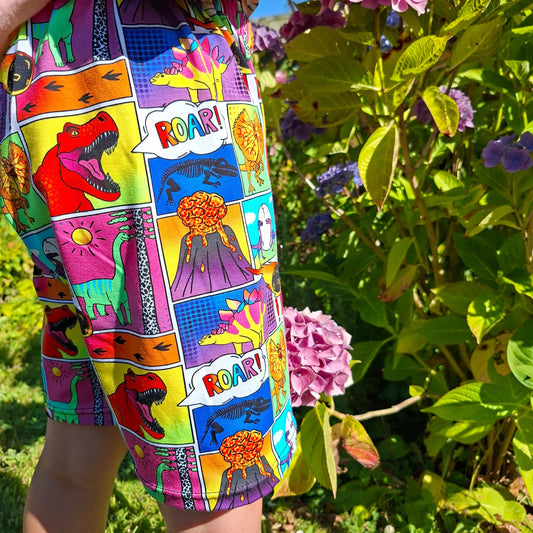 A close-up of a colourful children's shorts or skirt featuring a comic-book style dinosaur print, worn by a child outdoors in front of green hydrangea bushes.