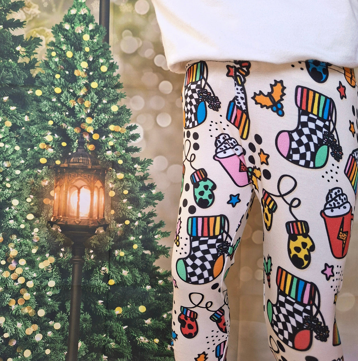 A child wearing white leggings with a colorful Christmas-themed print of stockings, hot air balloons, candy canes, and stars, standing in front of a decorated Christmas tree and a glowing street lamp.