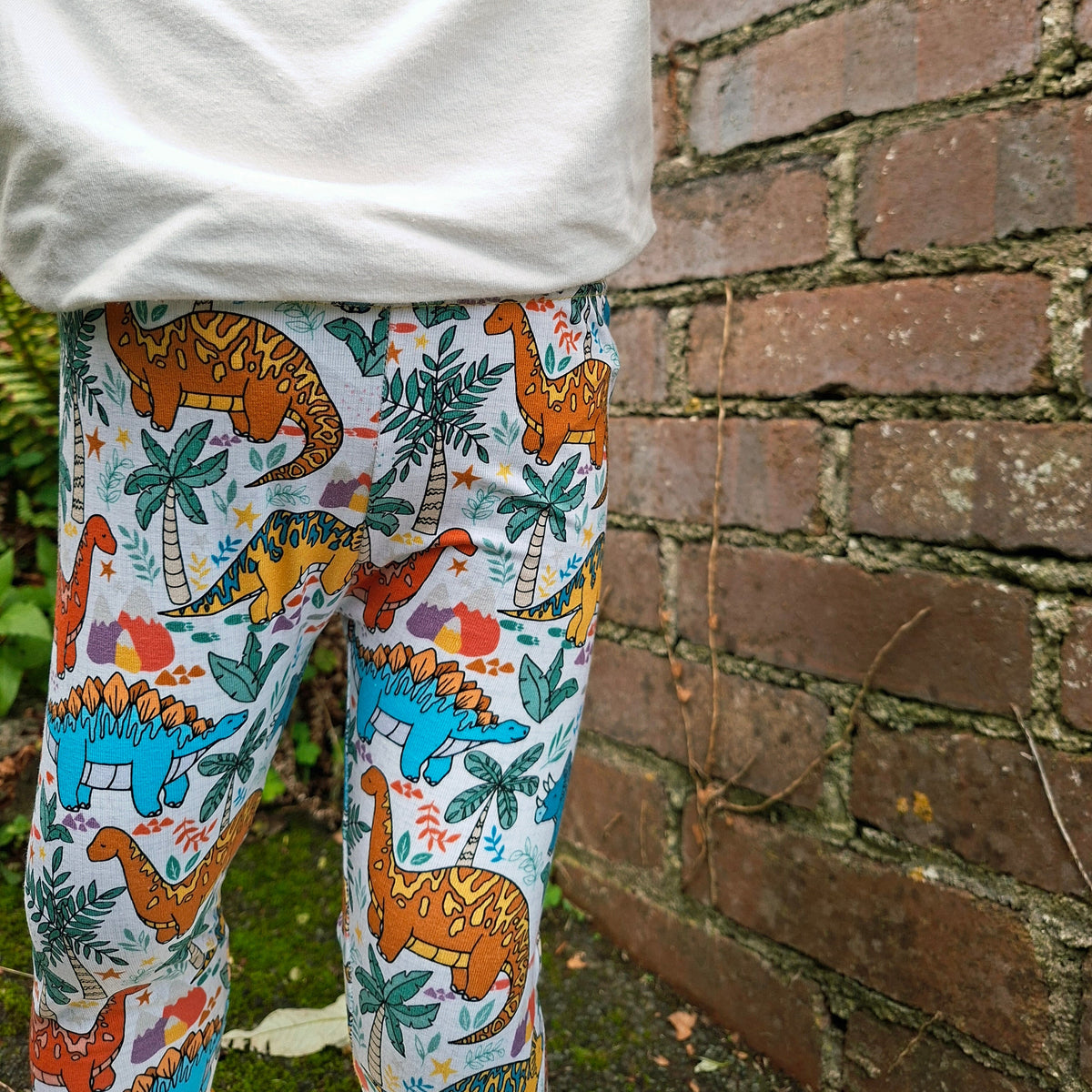 A child wearing colorful dinosaur-print leggings and a white sweatshirt, standing next to a brick wall outdoors.