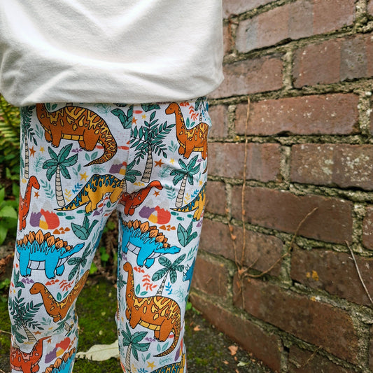 A child wearing colorful dinosaur-print leggings and a white sweatshirt, standing next to a brick wall outdoors.