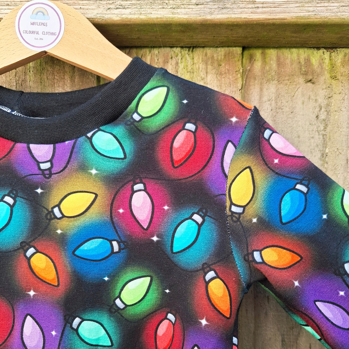 A long-sleeve crewneck twirly dress featuring a colourful Christmas light bulb pattern on a black background, hanging on a wooden hanger against a rustic wooden wall.