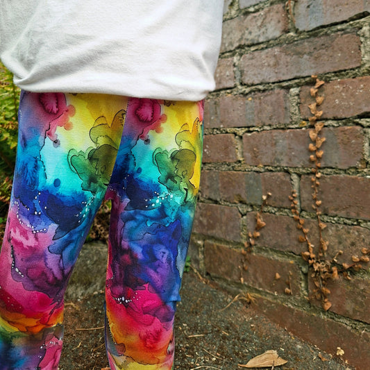 A child wearing colourful ink swirl watercolor-print leggings and a white long-sleeve top, standing outdoors against a brick wall.