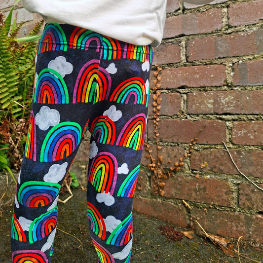A person wearing colourful rainbow and cloud print leggings with a white sweatshirt, standing outdoors against a brick wall.