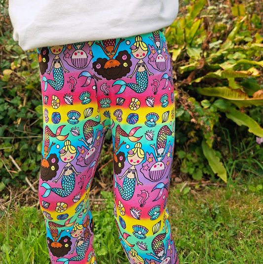 A child outdoors wearing colourful leggings with a mermaid and sea-themed print, paired with a white sweatshirt. The background shows green grass and plants.