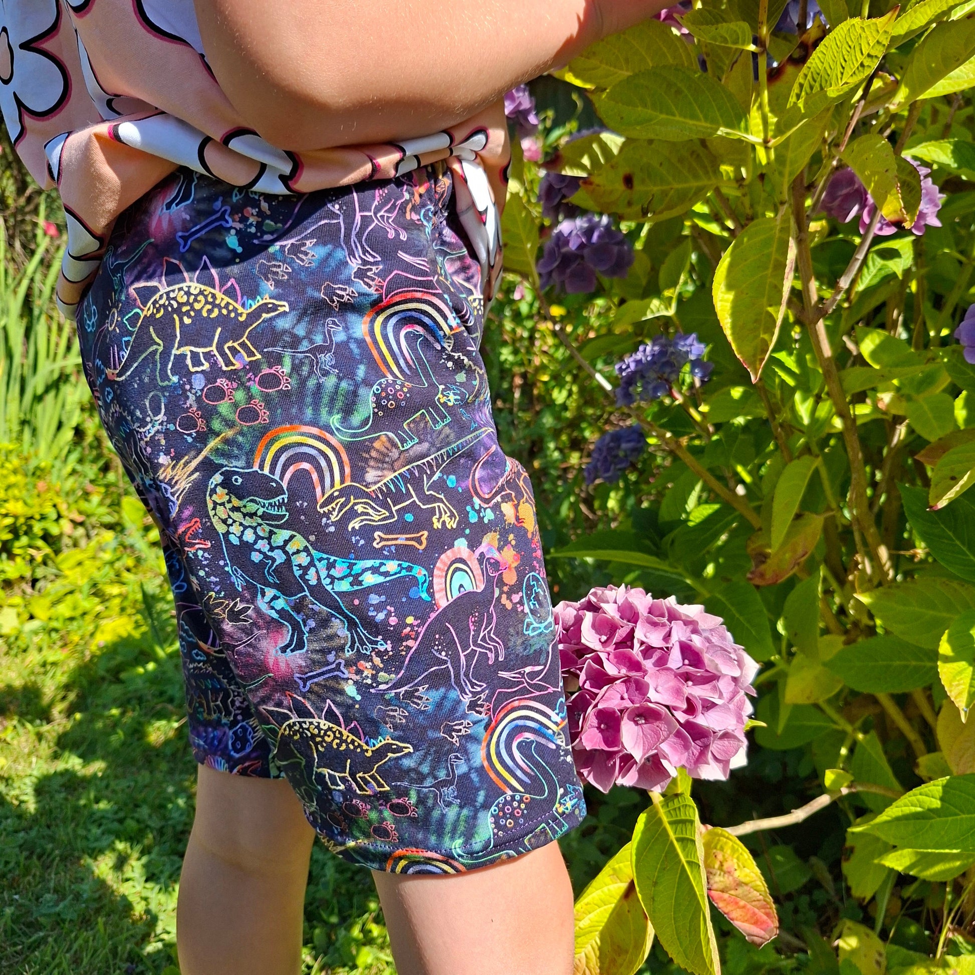 A child wearing colourful dinosaur print patterned shorts, photographed outdoors among green leaves and pink flowers.