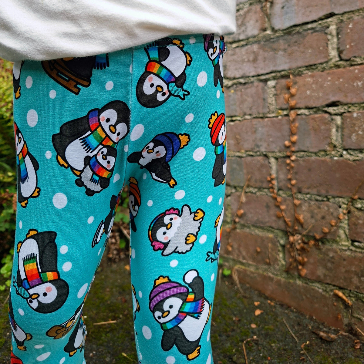 A child wearing bright turquoise leggings with a playful penguin print, paired with a white long-sleeve top, standing outdoors against a brick wall.
