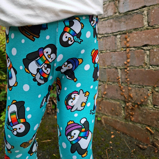 A child wearing bright turquoise leggings with a playful penguin print, paired with a white long-sleeve top, standing outdoors against a brick wall.