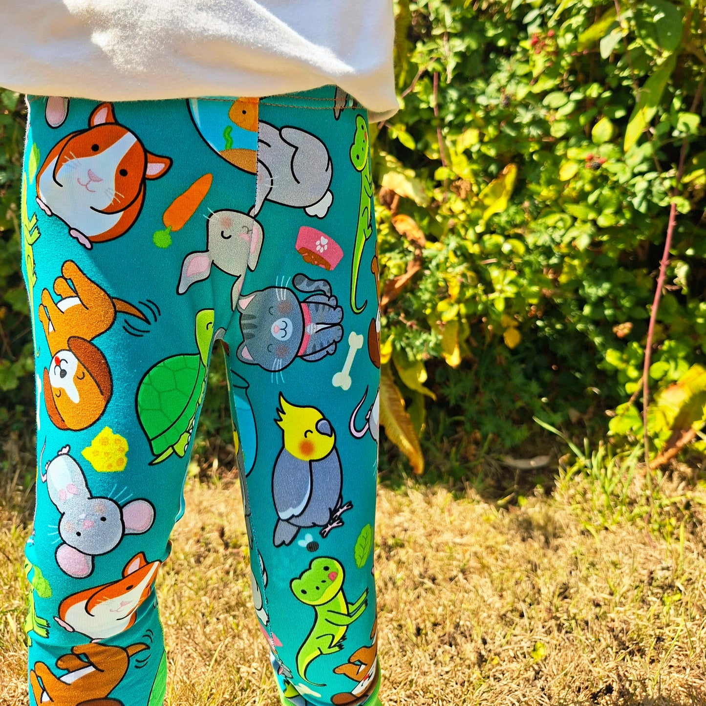 A close-up of a child's legs wearing bright turquoise leggings with a colorful cartoon animal print, standing outdoors in a grassy garden area.