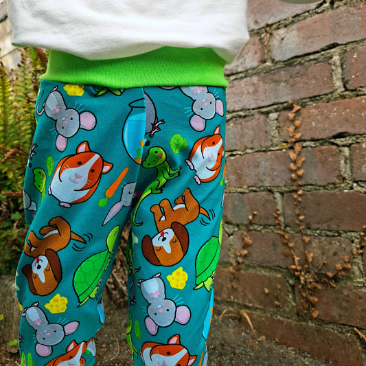 A child wearing bright turquoise relaxed fit style are with a colorful cartoon animal print, paired with a white top and green waistband, standing outdoors against a brick wall.