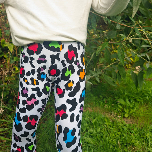 A child wearing white leggings with a colorful leopard print pattern, paired with a cream sweatshirt, standing outdoors in a grassy, leafy environment.