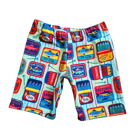 A pair of kids' shorts featuring a colorful cartoon sardine can print on a light blue striped background, displayed flat against a white background.