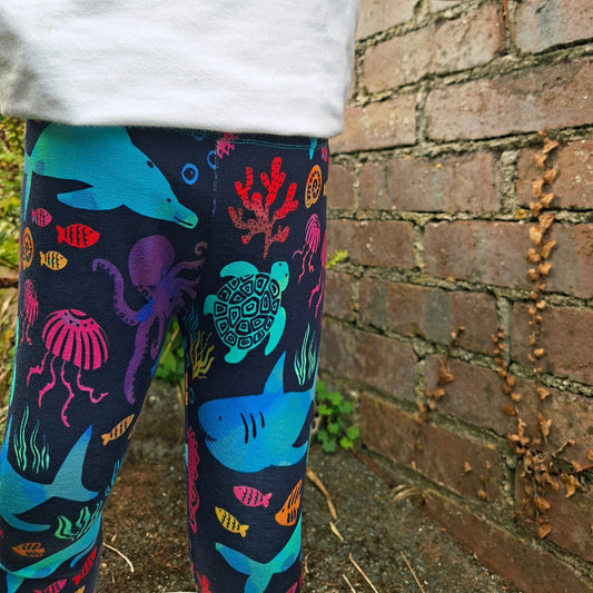 A child wearing colourful underwater-themed leggings with a white long-sleeve top, standing next to a brick wall.