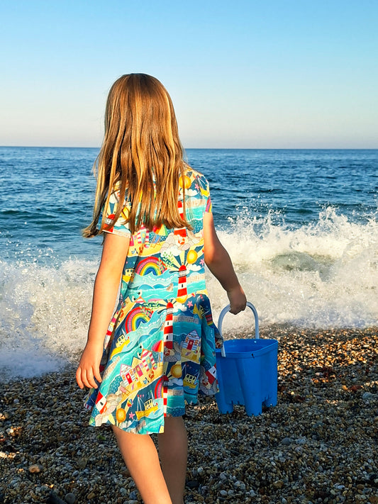 Child in a colorful dress standing on a beach with a blue bucket and sand toys.