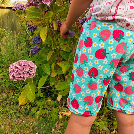 A child wearing bright turquoise shorts with a pink strawberry and white daisy pattern, standing outdoors next to purple hydrangea flowers.