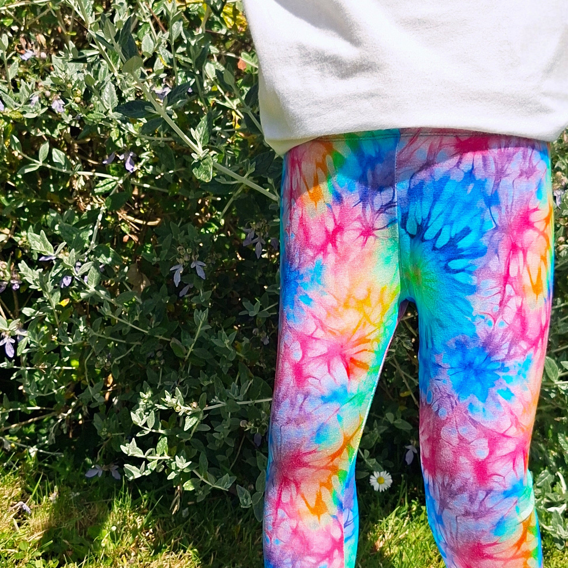 Child wearing colorful tie-dye leggings standing outdoors with greenery in the background