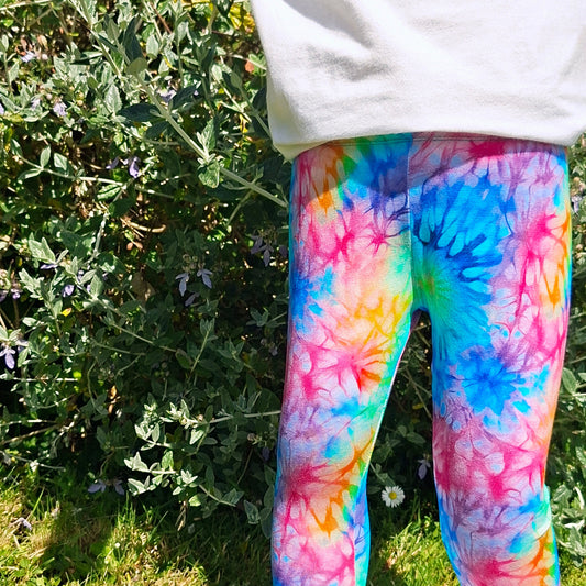 Child wearing colorful tie-dye leggings standing outdoors with greenery in the background