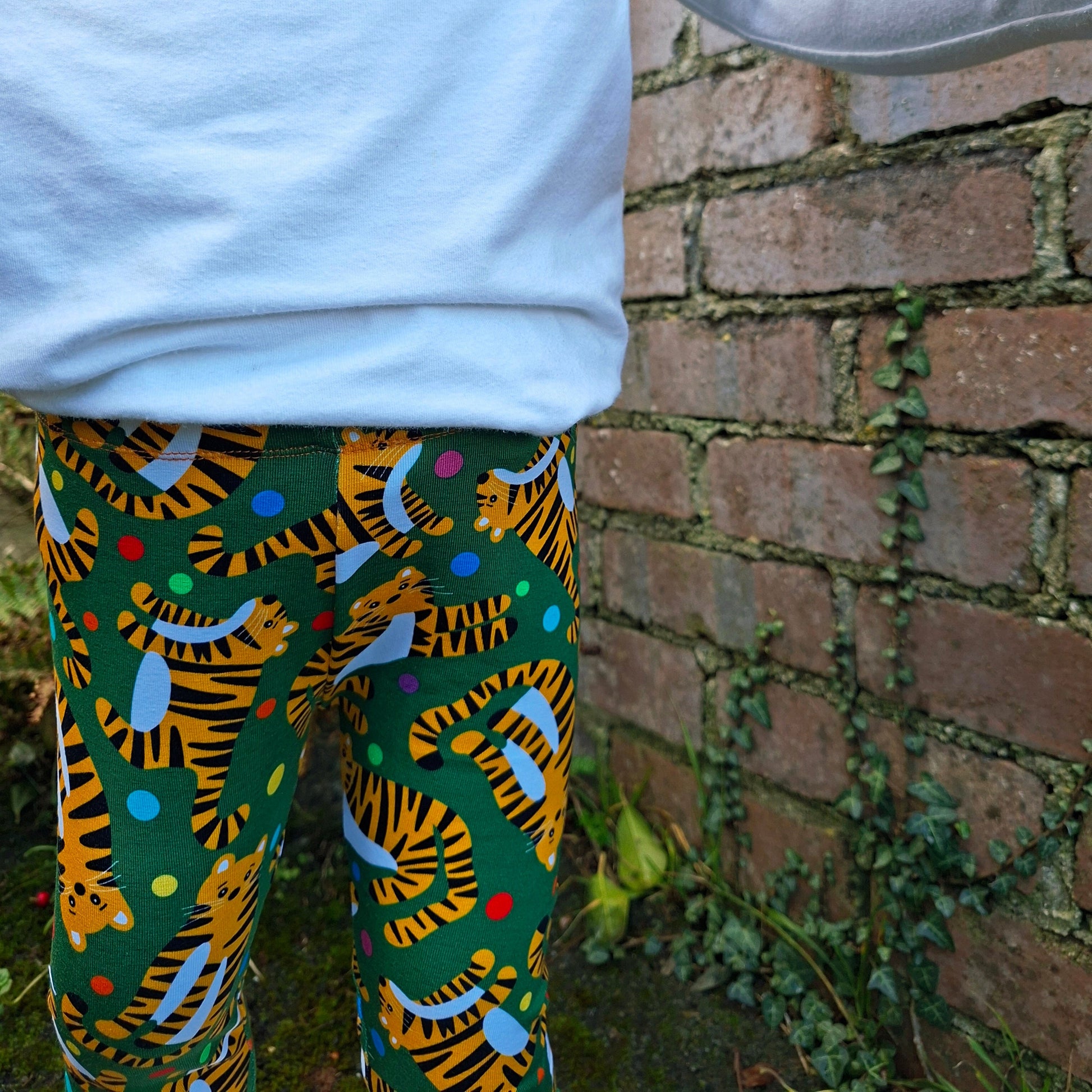 Child wearing colorful tiger-patterned pants against a brick wall.