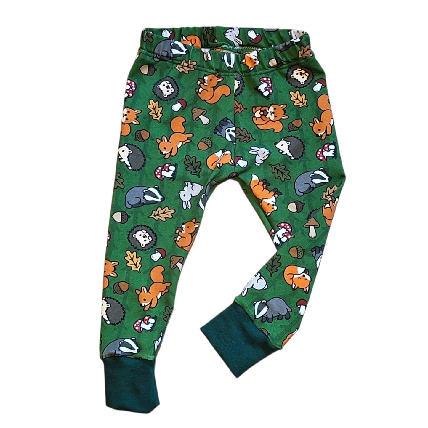 Green children's leggings with an all-over woodland animal print featuring foxes, hedgehogs, badgers, squirrels, acorns, and mushrooms, finished with solid dark green ribbed ankle cuffs.