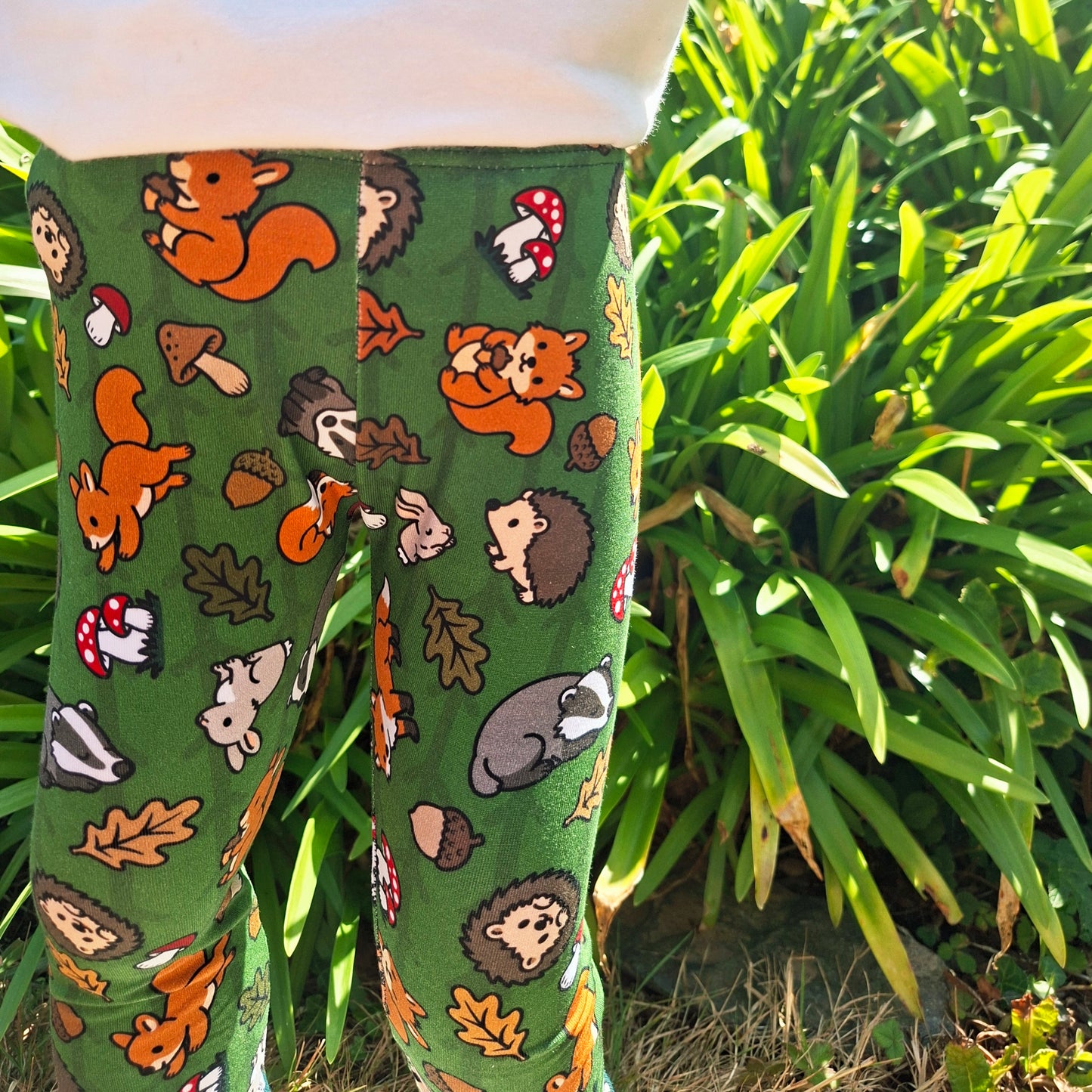 Green children's leggings with a colourful woodland animal print of squirrels, hedgehogs, acorns, and mushrooms, shown on a child outdoors in front of green plants.