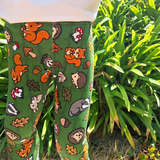 Green children's leggings with a colourful woodland animal print of squirrels, hedgehogs, acorns, and mushrooms, shown on a child outdoors in front of green plants.