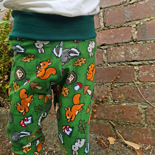 A child wearing green relaxed fit trousers with a woodland animal print of squirrels, badgers, hedgehogs, and acorns, paired with a white sweatshirt, standing in front of a brick wall.