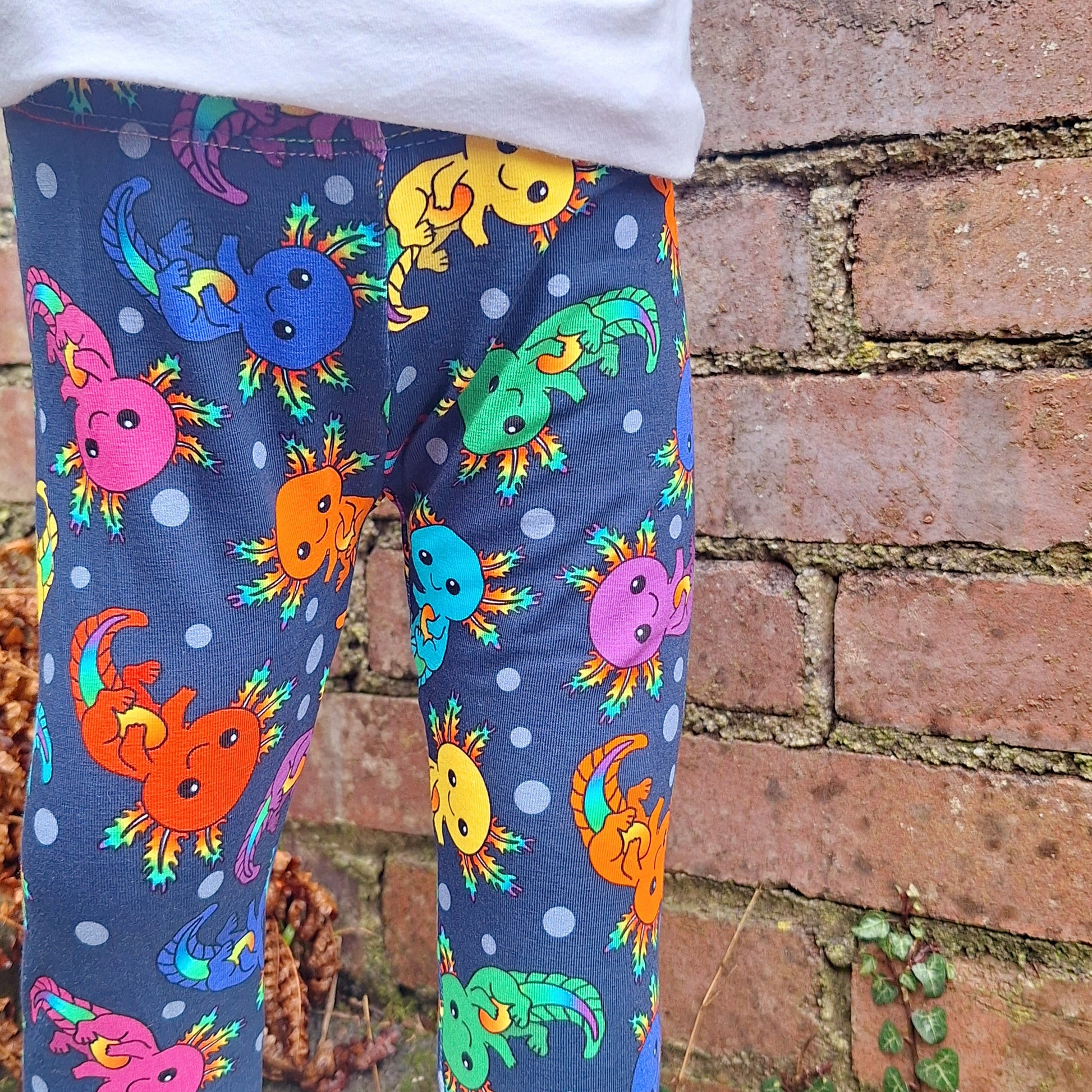 Child wearing multicolored axolotl patterned cuffed leggings with an elastic waistband, standing against a brick wall.
