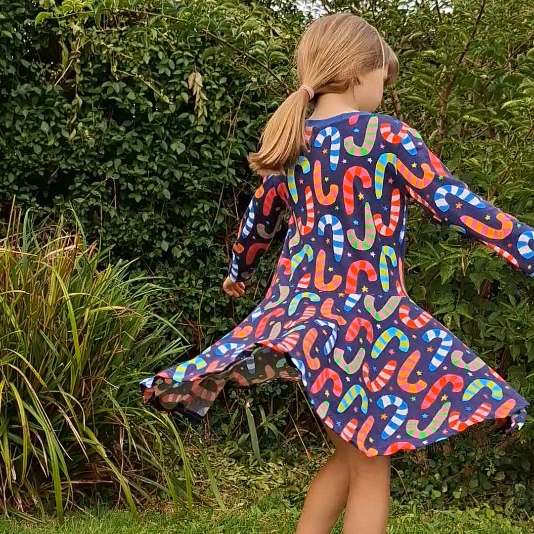 A young girl with blonde hair in a ponytail is seen from behind, wearing a long-sleeved blue dress with a colorful candy cane pattern, standing on a grassy lawn in front of green bushes.