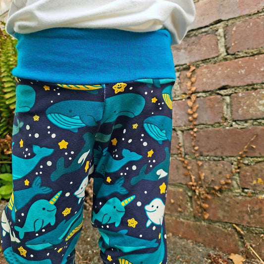 A child wearing patterned navy relaxed fit style trousers with a blue waistband, featuring a print of whales, narwhals, and yellow stars, standing outdoors against a brick wall.