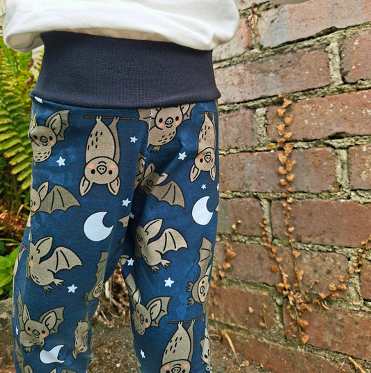 A child wearing blue relaxed fit trousers with a cartoon bat and moon pattern, paired with a white sweatshirt, standing against a brick wall.